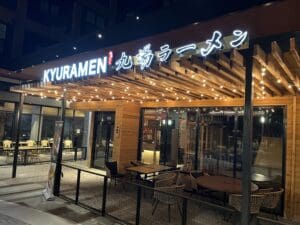 Exterior of KYURAMEN restaurant at night with illuminated sign, string lights, wooden pergola, tables, and chairs on the outdoor patio.