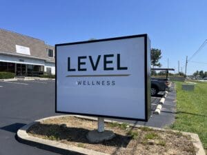 A large rectangular sign in a parking lot reads "LEVEL WELLNESS" in bold letters, with a building displaying the same name in the background.