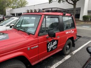 A red Mitsubishi Raider SUV with "Bolts Electrical Services" and "DONE MY JOB!" written on the side, parked in a lot near office buildings.