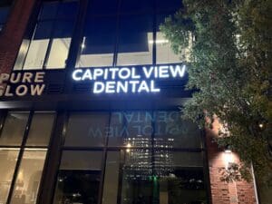 A brightly lit "Capitol View Dental" sign on the exterior of a building at night, with its reflection visible in the window below.