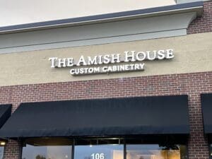 Storefront sign reads "The Amish House Custom Cabinetry" on a brick and tan building, with black awnings below.