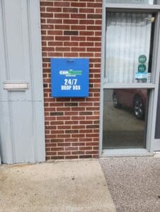 A blue 24/7 drop box is mounted on a brick wall next to a door with glass panels at the entrance of a building.