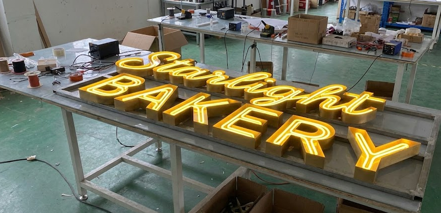 A lit "Starlight BAKERY" sign rests on a workbench in a workshop, surrounded by tools, wires, and cardboard boxes.