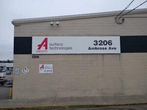 A beige building with signs reading "aalberts surface technologies" and the address "3206 Ambrose Ave." The entrance is marked with an additional sign and arrow.