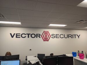 Office wall with the words "VECTOR SECURITY" and a red geometric logo, with several computer stations and office equipment visible in the foreground.