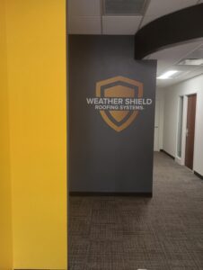Interior hallway with gray wall displaying the Weather Shield Roofing Systems logo in gold and white, yellow wall on the left, carpeted floor, and office doors in the background.