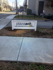 A white sign reads "plaza art Artist Materials & Picture Framing" on a patch of grass near a sidewalk, with buildings and a street in the background.