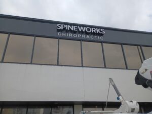 The exterior of a building with large windows features a sign that reads "Spineworks Chiropractic." A lift vehicle is partially visible in the lower right corner.