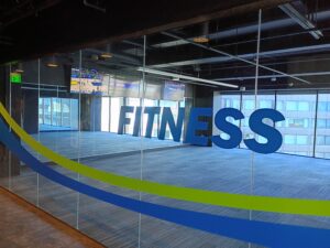 A spacious, empty fitness room with large windows, blue carpet, and the word "FITNESS" on the glass wall; TVs show sports in the background.