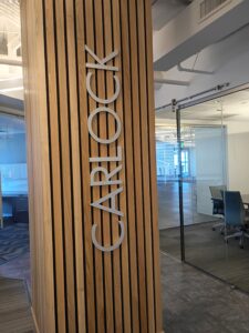 Wooden vertical slat column with the word "CARLOCK" in large silver letters; modern office space with glass walls and conference room in the background.