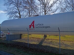 A large white industrial tank labeled "Aalberts surface technologies" stands behind a chain-link fence with trees and grass in the background.