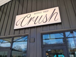 Sign reading "Crush Bottle Shop" mounted above glass doors on a dark-paneled building exterior. Reflections of trees and sky are visible in the windows.