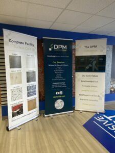 Three standing banners display information about DPM Surface Care, including services, core values, and surface care options, set in an office with blue walls and ceiling tiles.