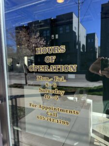 Glass door with gold lettering showing business hours: Mon-Fri 8-6, Sat 10-5, and appointment phone number 615-742-1799. Buildings and reflection visible in background.