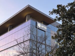 A modern glass office building with a BMG logo sign on the upper corner, reflecting trees and a blue sky at sunset.