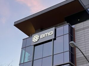 The exterior of a modern building with a prominent "BMG" sign and logo on the glass facade, seen from below under a partly cloudy sky.