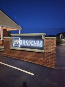 Illuminated sign for Knowles Private Wealth is mounted on a brick base outside a building at dusk.