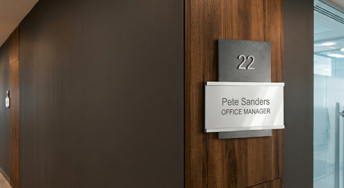Office door with a sign reading "22" and "Pete Sanders, Office Manager" mounted on a wooden wall; glass office interior visible in the background.