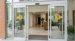 Automatic sliding glass doors open to a building entrance with caution signs, visible plants, and a reception desk inside.