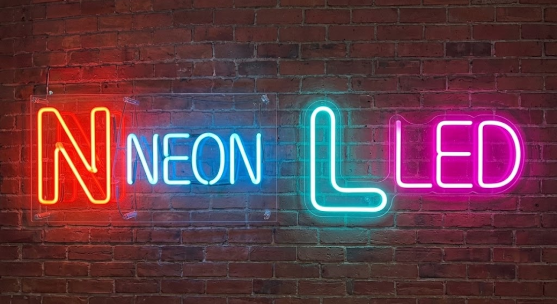 Neon sign on brick wall reads “NEON LED”; “N” and “L” letters are larger and outlined in different neon colors.